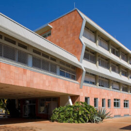 Das Bild zeigt ein modernes, vierstöckiges Gebäude mit einer Fassade aus rotem Backstein und weißen Metallelementen. Große Fenster mit horizontalen Jalousien prägen das Erscheinungsbild, vor dem Eingang befinden sich Pflanzen und Sträucher. Im Hintergrund sind mehrere geparkte Autos zu sehen, der Himmel ist klar und blau.