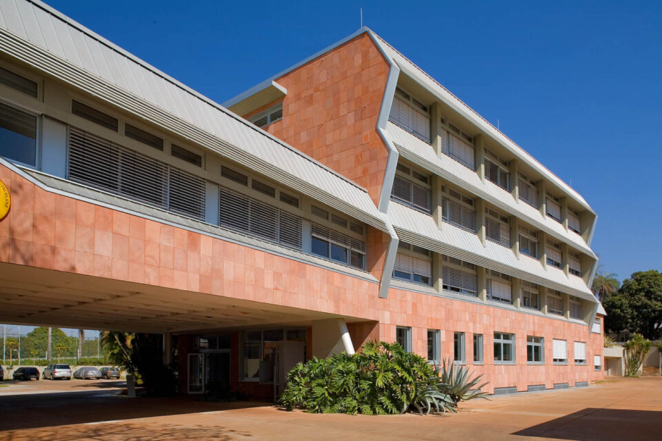Das Bild zeigt ein modernes, vierstöckiges Gebäude mit einer Fassade aus rotem Backstein und weißen Metallelementen. Große Fenster mit horizontalen Jalousien prägen das Erscheinungsbild, vor dem Eingang befinden sich Pflanzen und Sträucher. Im Hintergrund sind mehrere geparkte Autos zu sehen, der Himmel ist klar und blau.
