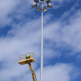Das Bild zeigt einen hohen Lichtmast mit mehreren Flutlichtstrahlern, die kreisförmig am oberen Ende angeordnet sind. Eine Person steht auf einer hydraulischen Hebebühne in luftiger Höhe und arbeitet in der Nähe des Mastes. Im Hintergrund ist ein teils bewölkter Himmel mit blauen Abschnitten zu sehen.