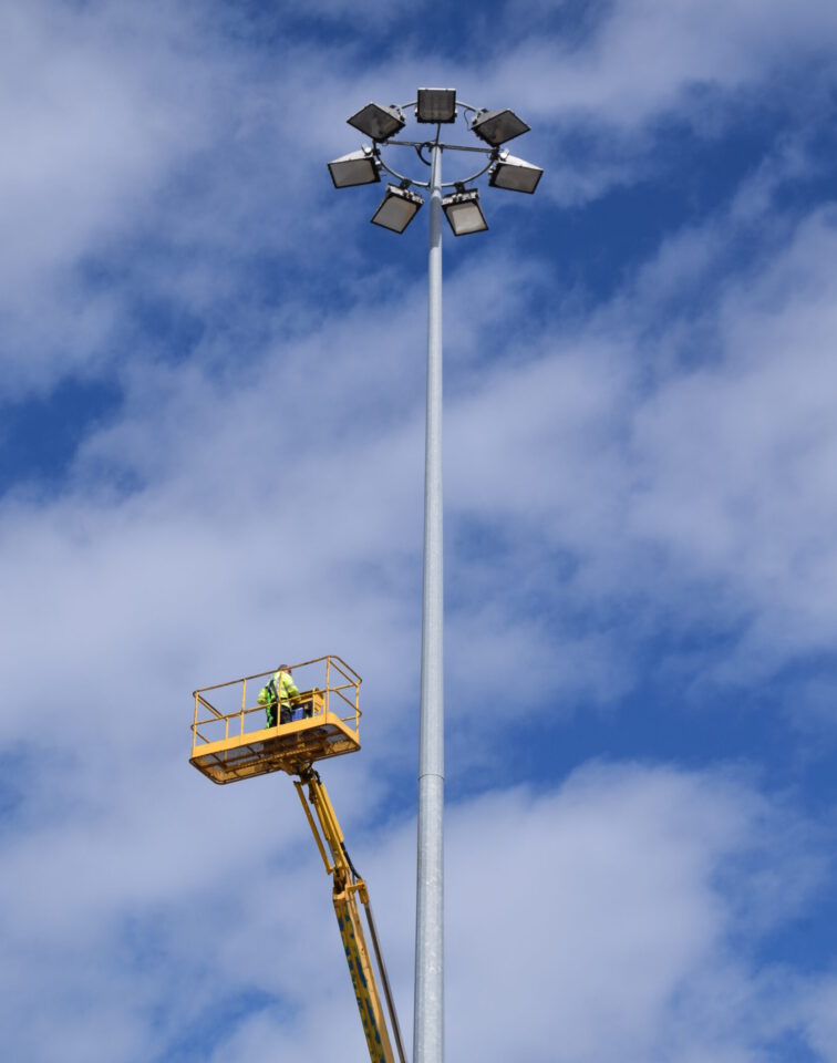 Das Bild zeigt einen hohen Lichtmast mit mehreren Flutlichtstrahlern, die kreisförmig am oberen Ende angeordnet sind. Eine Person steht auf einer hydraulischen Hebebühne in luftiger Höhe und arbeitet in der Nähe des Mastes. Im Hintergrund ist ein teils bewölkter Himmel mit blauen Abschnitten zu sehen.
