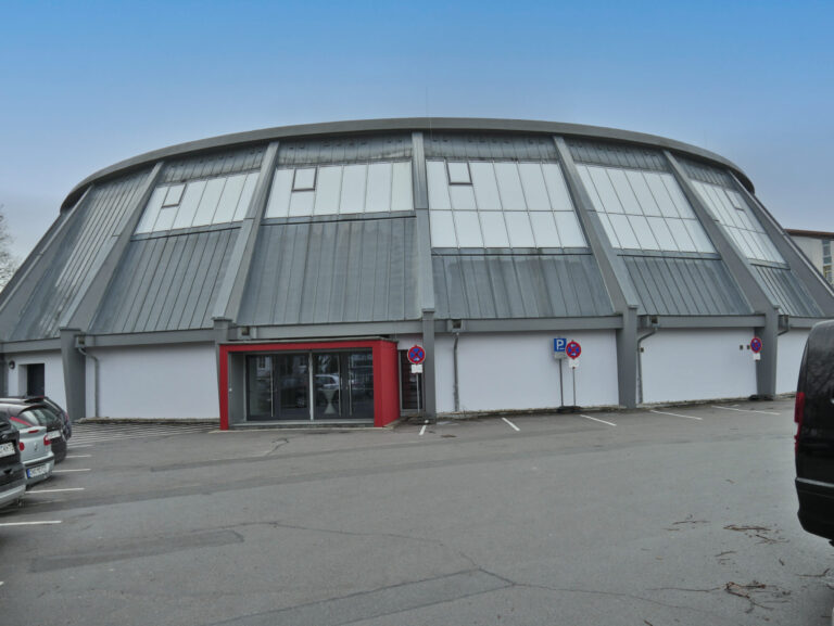 Das Bild zeigt ein großes, rundes Gebäude mit einer kuppelartigen Struktur, bestehend aus Metallpaneelen und Glasfenstern. In der Mitte befindet sich eine Eingangstür mit rotem Rahmen. Vor dem Gebäude sind mehrere Autos geparkt und es sind Parkschilder in der Nähe des Eingangs zu sehen.