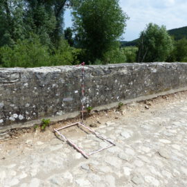 Das Bild zeigt eine Steinmauer, vor der ein rechteckiger Messrahmen aus rot-weiß gestreiften Stangen auf dem Kopfsteinpflaster liegt. Im Hintergrund sind grüne Bäume und Büsche zu sehen, was auf eine Außenaufnahme hinweist.