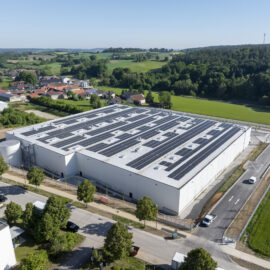 Das Bild zeigt eine Luftaufnahme eines großen Industriegebäudes mit Solarpaneelen auf dem Dach. Das Gebäude liegt in ländlicher Umgebung mit Straßen, grünen Feldern und einem kleinen Dorf im Hintergrund. Einige Fahrzeuge sind in der Nähe des Gebäudes geparkt.