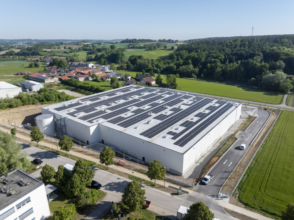 Das Bild zeigt eine Luftaufnahme eines großen Industriegebäudes mit Solarpaneelen auf dem Dach. Das Gebäude liegt in ländlicher Umgebung mit Straßen, grünen Feldern und einem kleinen Dorf im Hintergrund. Einige Fahrzeuge sind in der Nähe des Gebäudes geparkt.