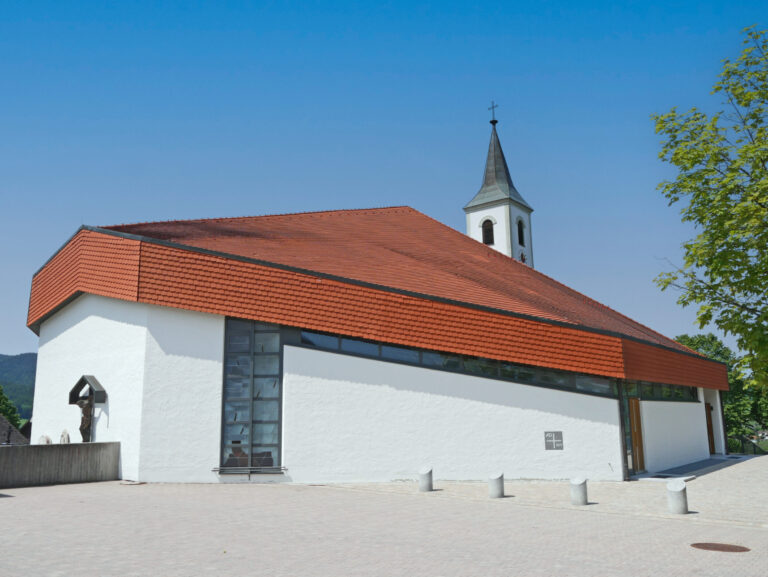 Das Bild zeigt ein modernes Kirchengebäude mit weißer Fassade und rotem Ziegeldach. Die Architektur ist kantig gestaltet, mit großen Fenstern entlang einer Seite. Im hinteren Bereich befindet sich ein Glockenturm mit einem Kreuz an der Spitze, davor ein gepflasterter Platz mit mehreren Pollern.