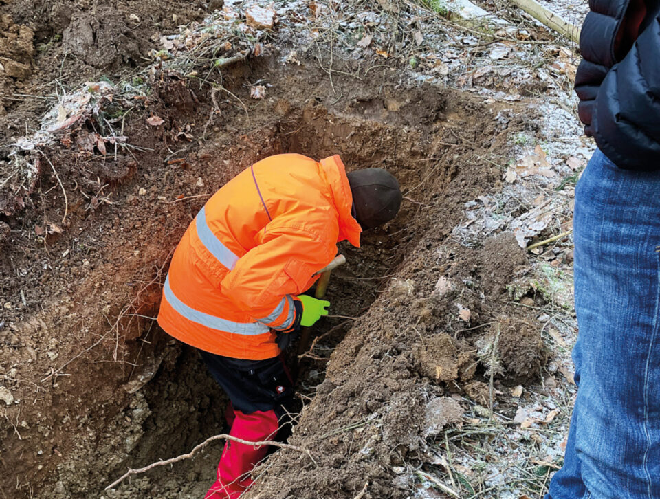 Das Bild zeigt eine Person in orangefarbener Warnkleidung mit reflektierenden Streifen, grünen Handschuhen und roten Stiefeln, die in einem Erdloch oder Graben steht und dort arbeitet oder etwas untersucht. Rechts am Bildrand ist eine weitere Person teilweise zu sehen. Der Boden ist teilweise mit Frost oder Schnee bedeckt und von natürlicher Vegetation umgeben.