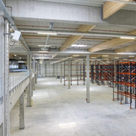 Das Bild zeigt einen Weg aus hellen Pflastersteinen entlang einer Gebäudewand aus Metall. Links am Weg wachsen junge Weinreben an Drahtgestellen, eine davon ist mit einem Schild mit der Aufschrift „Weintraube“ und einem Traubenbild versehen. Rechts des Weges befindet sich ein trockener Hang mit spärlicher Vegetation und einer Betonmauer am oberen Ende.