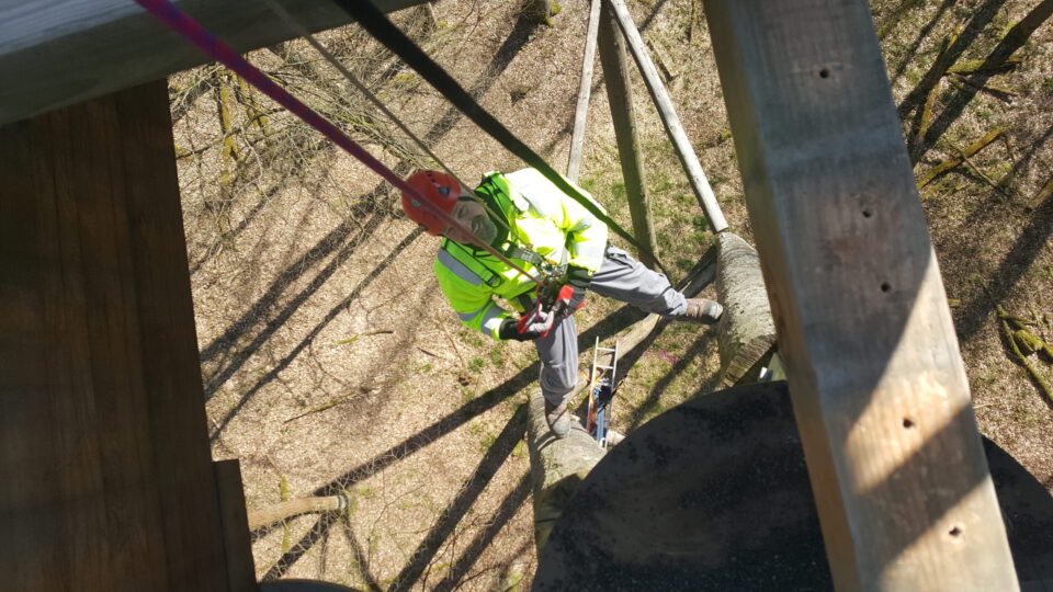 Das Bild zeigt eine Person mit Warnweste, Helm und Sicherheitsgurt, die in luftiger Höhe an einer Holzkonstruktion arbeitet. Die Person hält sich an Seilen fest und scheint Wartungs- oder Bauarbeiten durchzuführen. Im Hintergrund sind Bäume und Schatten auf dem Boden zu erkennen.
