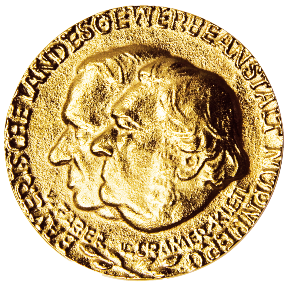 Das Bild zeigt eine goldfarbene Medaille mit den geprägten Profilen zweier Köpfe in der Mitte. Am Rand der Münze verläuft eine Inschrift in Großbuchstaben.