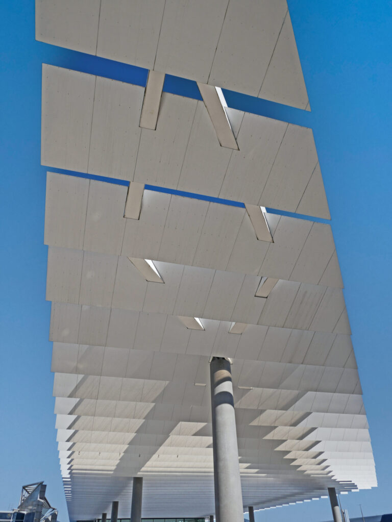 Das Bild zeigt eine große Konstruktion aus mehreren rechteckigen Paneelen, die in einem gitterartigen Muster angeordnet sind. Die Paneele werden von senkrechten Säulen getragen und gehören vermutlich zu einer architektonischen oder technischen Anlage, möglicherweise zur Beschattung oder zur Nutzung von Solarenergie. Im Hintergrund ist ein klarer blauer Himmel zu sehen.