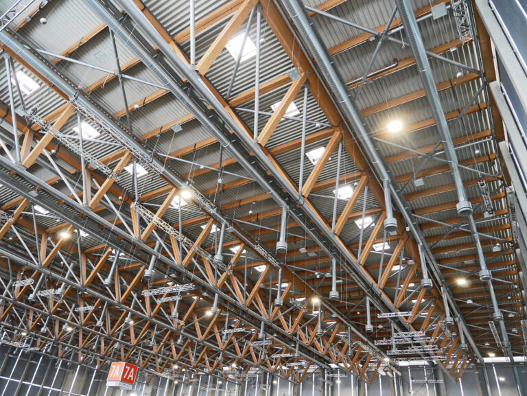 Das Bild zeigt die Decke eines großen Industrie- oder Gewerbegebäudes mit einem komplexen Geflecht aus Metall- und Holzträgern sowie Stützkonstruktionen. An der Decke sind mehrere Leuchten und Lüftungsrohre befestigt. Im Hintergrund sind rote Schilder mit der Aufschrift „7A“ auf weißem Grund zu sehen.