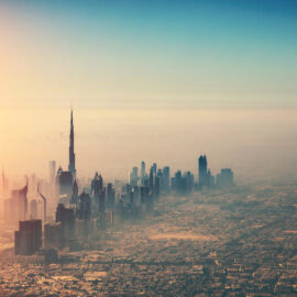 Das Bild zeigt eine Luftaufnahme einer Stadt mit zahlreichen Hochhäusern, im Zentrum ragt der Burj Khalifa als höchstes Gebäude hervor. Die Sonne geht links am Horizont auf oder unter und taucht die Stadt in ein warmes Licht. Der Himmel ist klar mit einem Farbverlauf von Blau zu Orange.