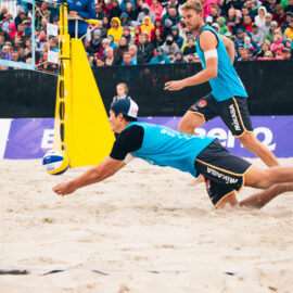 Das Bild zeigt ein Beachvolleyballspiel mit zwei Spielern im Vordergrund, beide in blauen Trikots und schwarzen Shorts. Einer der Spieler hechtet mit ausgestrecktem Arm nach dem Ball, während der andere im Hintergrund steht und bereit ist zu reagieren. Im Hintergrund sind ein gelber Netzpfosten, Zuschauertribünen und Werbebanner zu sehen.