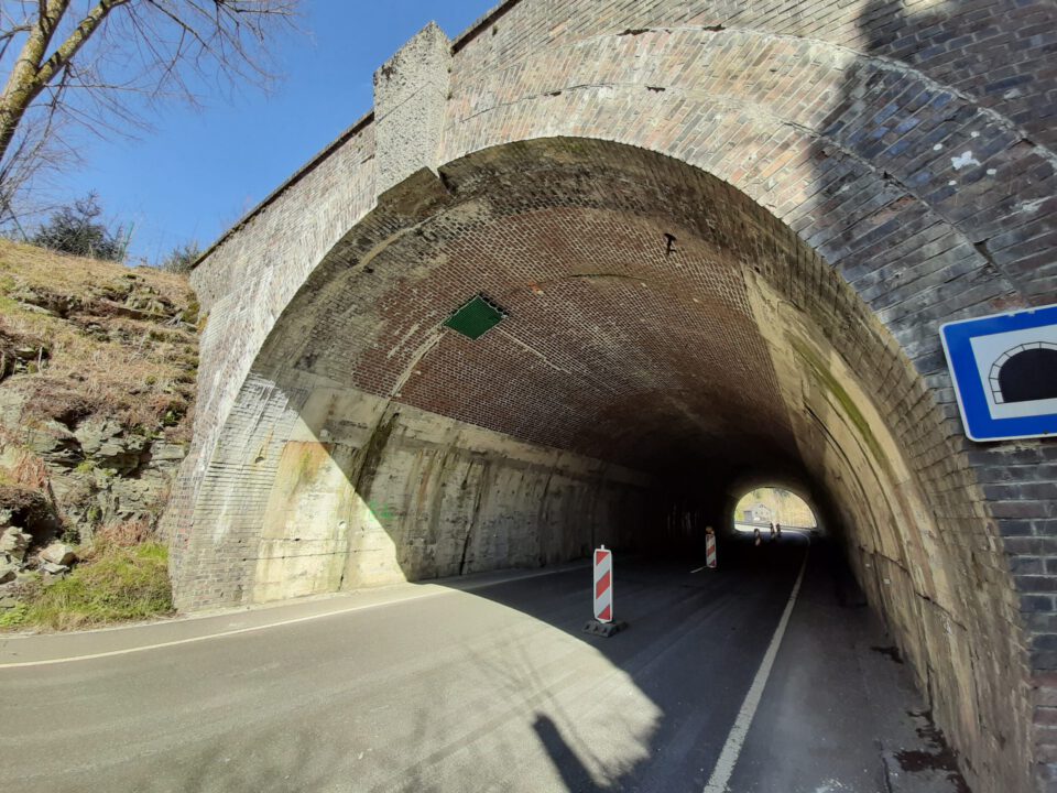 Das Bild zeigt eine Straße, die durch einen gemauerten Tunnel mit gewölbtem Deckengewölbe führt. Im Inneren des Tunnels stehen zwei Absperrbaken, die auf Bau- oder Wartungsarbeiten hinweisen. Die Umgebung außerhalb des Tunnels besteht aus felsigem Gelände mit etwas Vegetation und Bäumen auf der linken Seite.