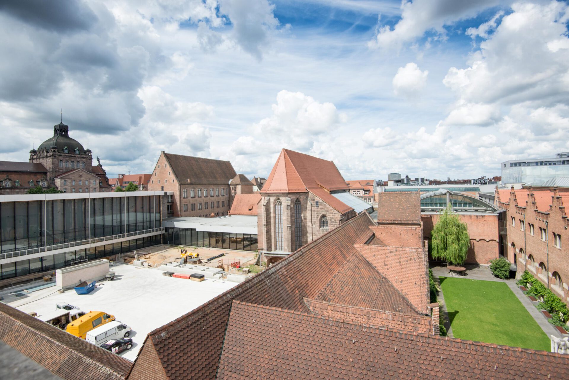 Das Bild zeigt eine historische Gebäudeanlage mit roten Ziegeldächern aus der Vogelperspektive. In der Mitte befindet sich ein Innenhof mit grünem Rasen und Bäumen, umgeben von modernen Glasbauten. Der Himmel ist teils bewölkt mit blauen Abschnitten.