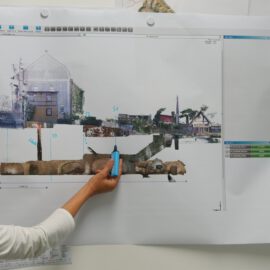 Das Bild zeigt eine Person, die mit einem blauen Textmarker auf ein großes, an der Wand angebrachtes Diagramm oder eine Karte zeigt. Die Darstellung zeigt einen Querschnitt eines städtischen Gebiets mit Gebäuden und unterirdischen Strukturen, ergänzt durch Markierungen und Maßangaben. Am oberen und rechten Rand sind Teile einer Software-Oberfläche zu sehen.