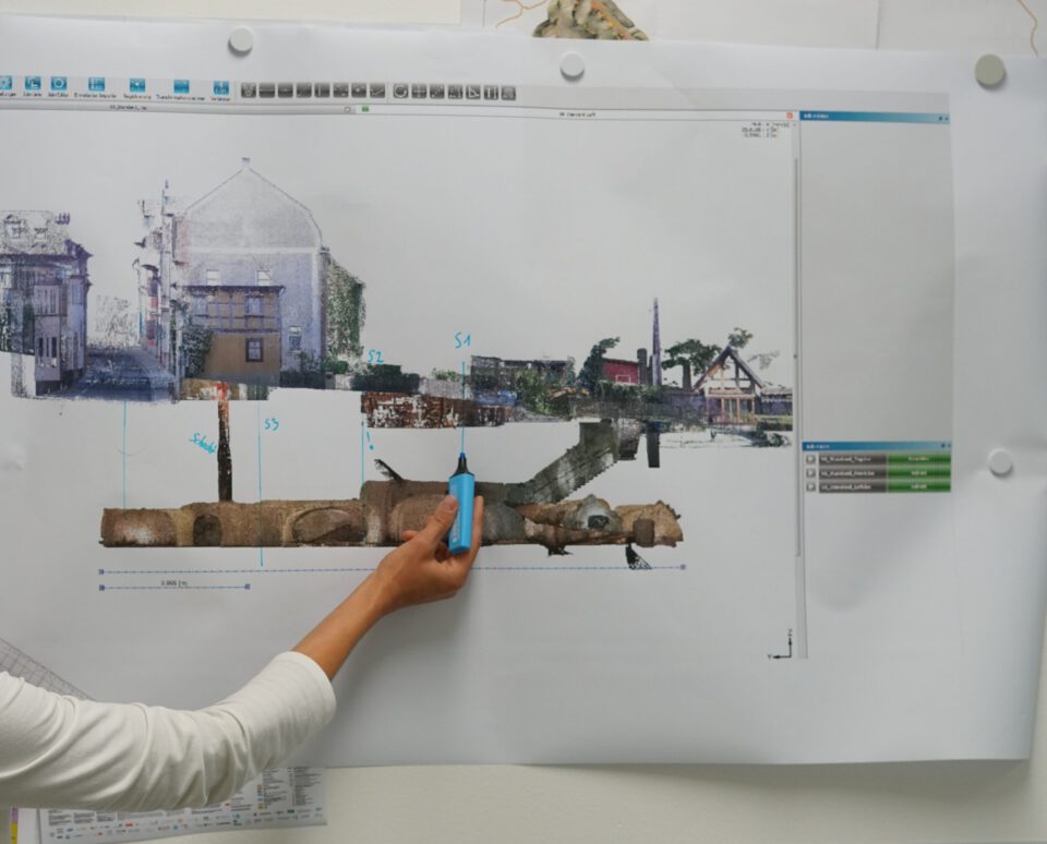 Das Bild zeigt eine Person, die mit einem blauen Textmarker auf ein großes, an der Wand angebrachtes Diagramm oder eine Karte zeigt. Die Darstellung zeigt einen Querschnitt eines städtischen Gebiets mit Gebäuden und unterirdischen Strukturen, ergänzt durch Markierungen und Maßangaben. Am oberen und rechten Rand sind Teile einer Software-Oberfläche zu sehen.