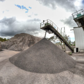 Das Bild zeigt ein Industriegelände mit einem großen Kies- oder Steinhaufen in der Mitte. Zwei erhöhte Förderbänder, gestützt von Metallkonstruktionen, verlaufen links und rechts und dienen dem Materialtransport. Im Hintergrund ist ein großer Erd- oder Felshügel zu sehen unter einem teils bewölkten Himmel.