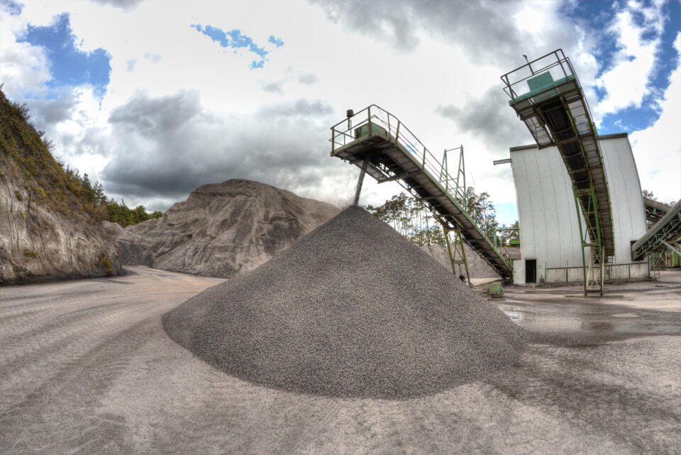 Das Bild zeigt ein Industriegelände mit einem großen Kies- oder Steinhaufen in der Mitte. Zwei erhöhte Förderbänder, gestützt von Metallkonstruktionen, verlaufen links und rechts und dienen dem Materialtransport. Im Hintergrund ist ein großer Erd- oder Felshügel zu sehen unter einem teils bewölkten Himmel.