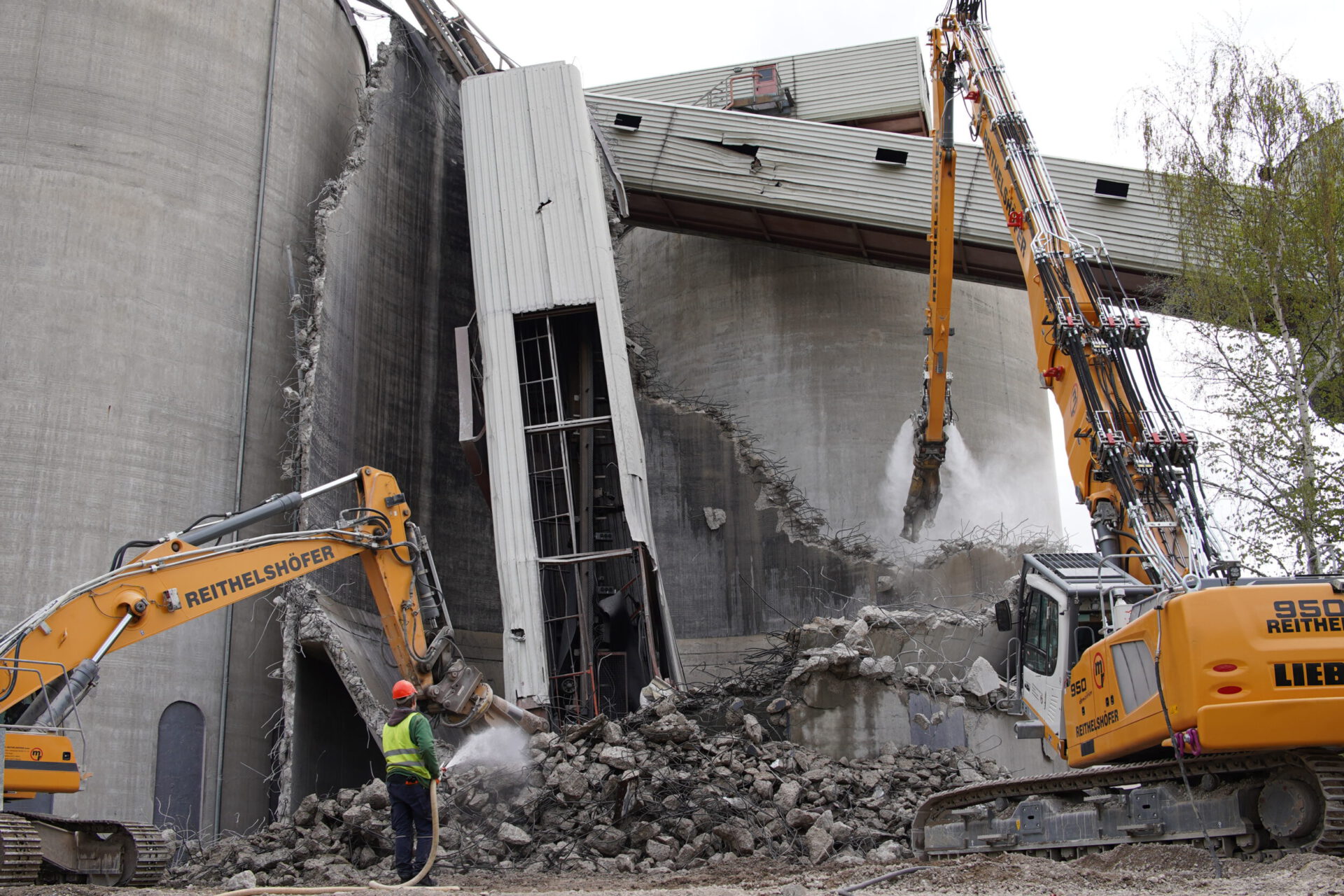 Auf dem Bild ist eine Abbruchstelle zu sehen, auf der zwei Bagger ein großes Betonbauwerk niederreißen. Ein Arbeiter mit grünem Schutzanzug und rotem Helm sprüht Wasser auf die Trümmer, um Staub zu binden. Die Szene zeigt eine fortgeschrittene Phase des Rückbaus mit deutlich sichtbaren Schäden am Bauwerk.