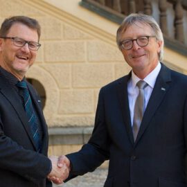 Das Bild zeigt zwei Personen in formeller Kleidung, die sich die Hand geben. Im Hintergrund ist ein Gebäude mit einer Steintreppe und einem Geländer zu sehen.