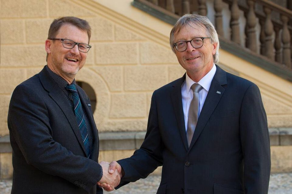Das Bild zeigt zwei Personen in formeller Kleidung, die sich die Hand geben. Im Hintergrund ist ein Gebäude mit einer Steintreppe und einem Geländer zu sehen.
