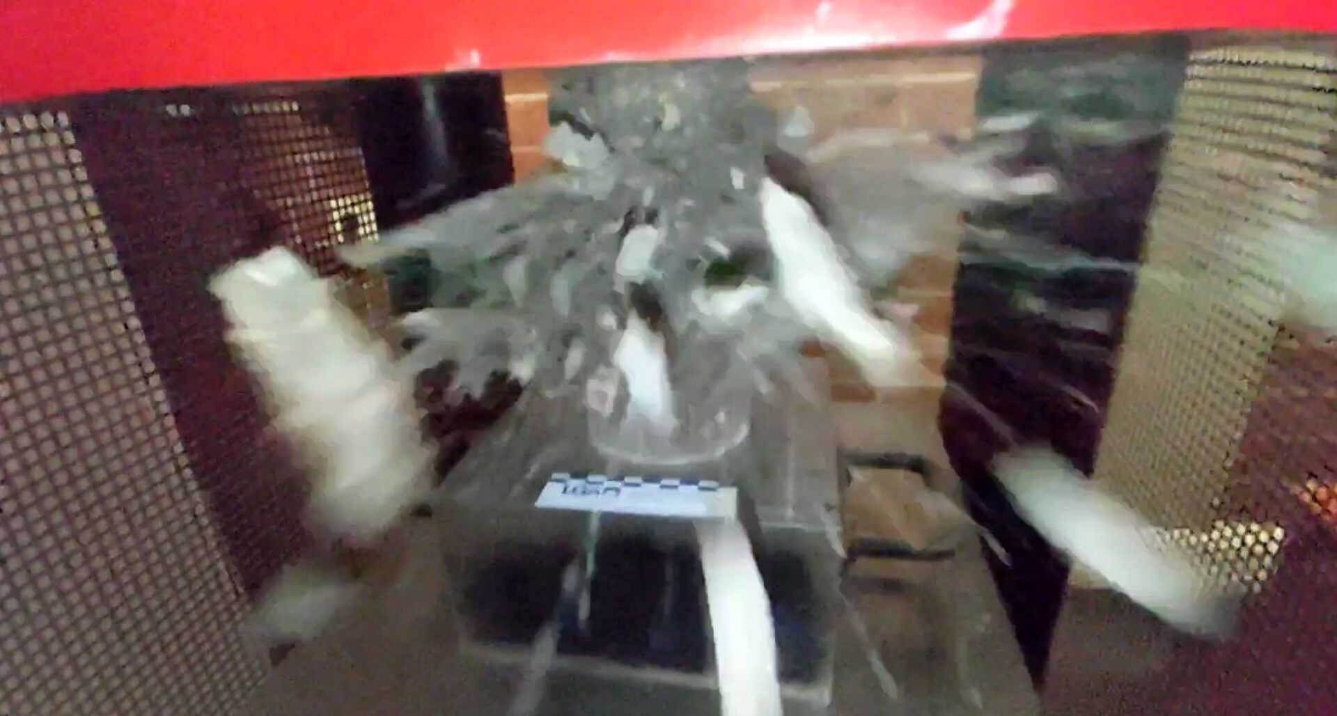 Das Bild zeigt eine Betonspezimen, die in einer kontrollierten Umgebung einem Festigkeitstest unterzogen wird. Der Beton zerbricht unter Druck, wobei Bruchstücke nach außen fliegen.