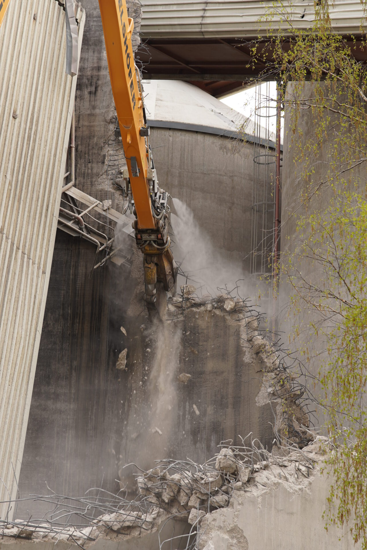 Auf dem Bild ist eine Baustelle zu sehen, auf der ein großer Bagger mit einem Hydraulikhammer eine Betonwand abbricht. Betonstücke und Staub fliegen durch die Luft, während Bewehrungsstahl sichtbar wird. Im Hintergrund sind weitere Industrieanlagen und Gerüste zu erkennen.