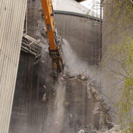 Auf dem Bild ist eine Baustelle zu sehen, auf der ein großer Bagger mit einem Hydraulikhammer eine Betonwand abbricht. Betonstücke und Staub fliegen durch die Luft, während Bewehrungsstahl sichtbar wird. Im Hintergrund sind weitere Industrieanlagen und Gerüste zu erkennen.