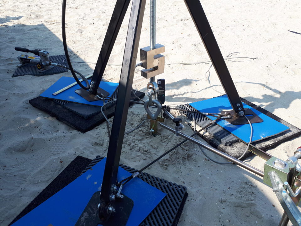 Das Bild zeigt eine Versuchsanordnung auf sandigem Untergrund mit mehreren Metallstrukturen. Drei schwarze Metallbeine bilden ein Stativ, das auf blauen Platten mit schwarzen Matten steht. In der Mitte hängt eine große metallene Messvorrichtung, welche mit Kabeln und Stangen verbunden ist.