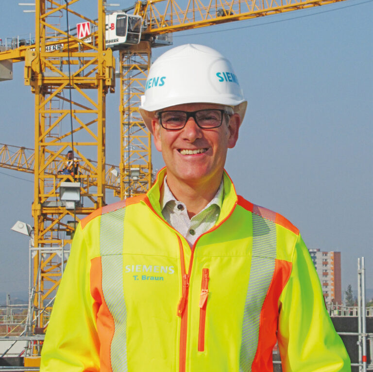 Das Bild zeigt eine Person mit weißem Schutzhelm und einer Warnweste, beide mit dem Siemens-Logo versehen. Im Hintergrund befinden sich große gelbe Baukräne und mehrere Gebäude.