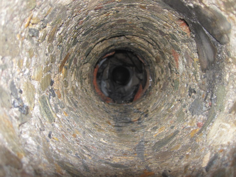 Das Bild zeigt das Innere einer zylindrischen Struktur mit unebenen Wänden aus Stein oder Beton. Die Perspektive führt in die Tiefe des Tunnels oder Rohrs, dessen Ende dunkel und weit entfernt erscheint. Die Oberfläche wirkt rau und gealtert.