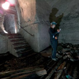 Das Bild zeigt eine Person mit Schutzkleidung und Helm mit Stirnlampe in einem alten, möglicherweise verlassenen Gebäude. Die Person hält ein Gerät in der Hand und scheint etwas zu untersuchen oder zu dokumentieren. Links führt eine Treppe zu einem gemauerten Torbogen, der Raum ist dunkel und von bröckelnden Stein- oder Betonwänden umgeben.