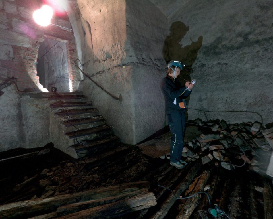 Das Bild zeigt eine Person mit Schutzkleidung und Helm mit Stirnlampe in einem alten, möglicherweise verlassenen Gebäude. Die Person hält ein Gerät in der Hand und scheint etwas zu untersuchen oder zu dokumentieren. Links führt eine Treppe zu einem gemauerten Torbogen, der Raum ist dunkel und von bröckelnden Stein- oder Betonwänden umgeben.
