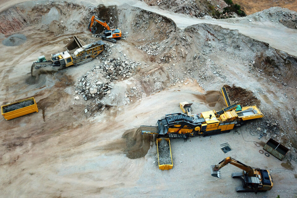Das Bild zeigt eine Luftaufnahme eines Steinbruchs oder Bergbaugeländes. Zwei große Maschinen, vermutlich Brecher oder Siebanlagen, verarbeiten das geförderte Material. Jeweils ein orangefarbener Bagger belädt die Maschinen, daneben stehen gelbe Container mit verarbeitetem Material.