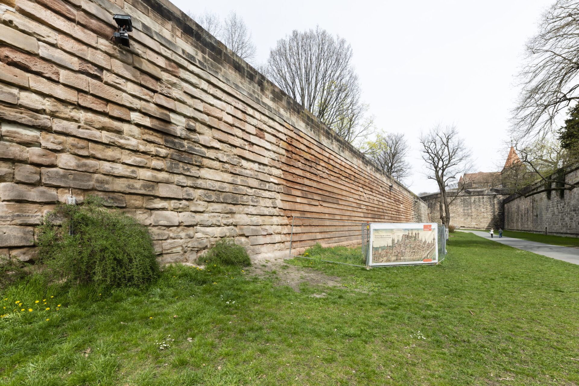 Das Bild zeigt eine große Steinmauer mit horizontal geschichteten Steinen. Davor befindet sich eine Grasfläche mit einigen Büschen und kleinen gelben Blumen sowie eine Informationstafel, die vermutlich historische oder erklärende Inhalte zeigt. Im Hintergrund sind kahle Bäume und ein Weg zu sehen, der in einen anderen Bereich führt, unter einem bewölkten Himmel.