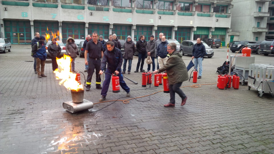 Das Bild zeigt eine Gruppe von Personen bei einer Feuerlöschübung im Freien. Zwei Personen bedienen aktiv Feuerlöscher, um ein kontrolliertes Feuer zu löschen, während weitere Teilnehmende im Hintergrund zuschauen. Im Hintergrund ist ein Gebäude mit nummerierten Toren zu sehen.