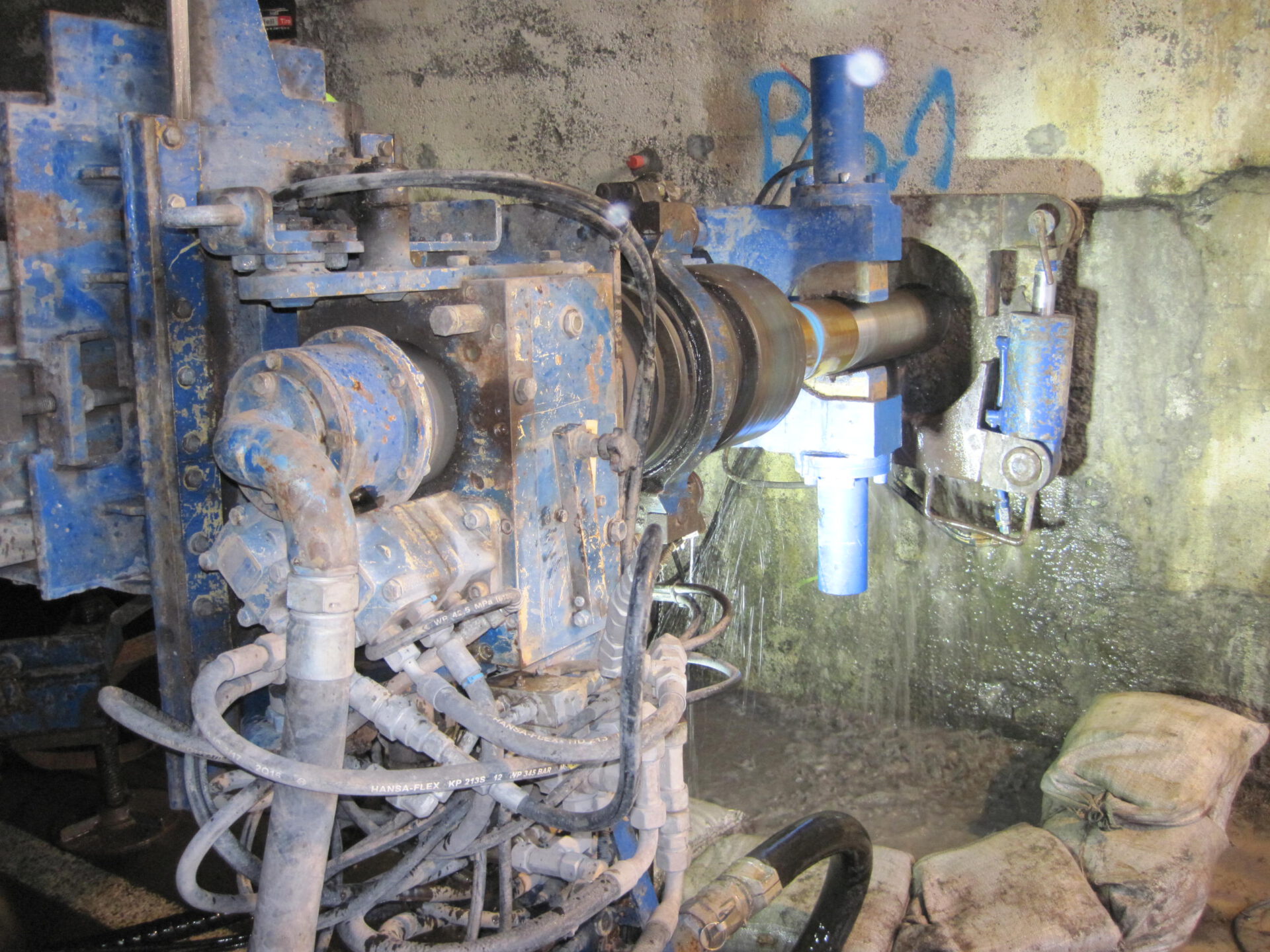 Das Bild zeigt eine schwere Industriemaschine, vermutlich für Bohr- oder Fräsarbeiten an einer Betonwand. Die Maschine ist blau und mit mehreren Hydraulikschläuchen und technischen Bauteilen ausgestattet. Sie befindet sich in einem Arbeitsumfeld mit Betonoberflächen.