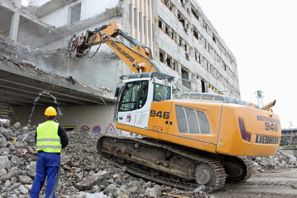 Das Bild zeigt eine Baustelle, auf der ein großer Bagger ein Gebäude abreißt. Der Bagger ist mit einem hydraulischen Abbruchhammer ausgestattet, der Betonteile des Gebäudes zerkleinert. Ein Bauarbeiter mit gelbem Helm und Warnweste steht in der Nähe und beobachtet den Abbruch.