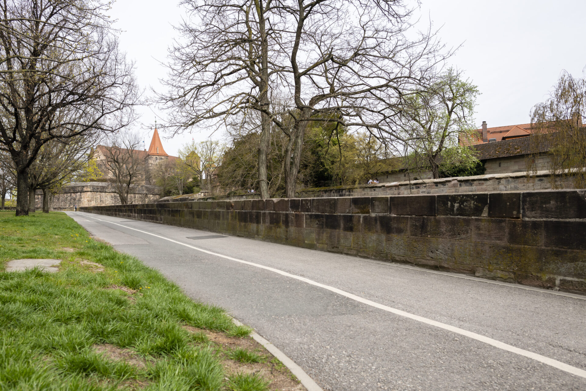 Das Bild zeigt eine asphaltierte Straße mit weißen Fahrbahnmarkierungen, die an einer Steinmauer entlangführt. Auf beiden Seiten der Straße stehen Bäume, teils mit, teils ohne Laub. Im Hintergrund ist ein Gebäude mit rotem Dach und spitzem Turm zu sehen, links neben der Straße befindet sich eine Grasfläche.