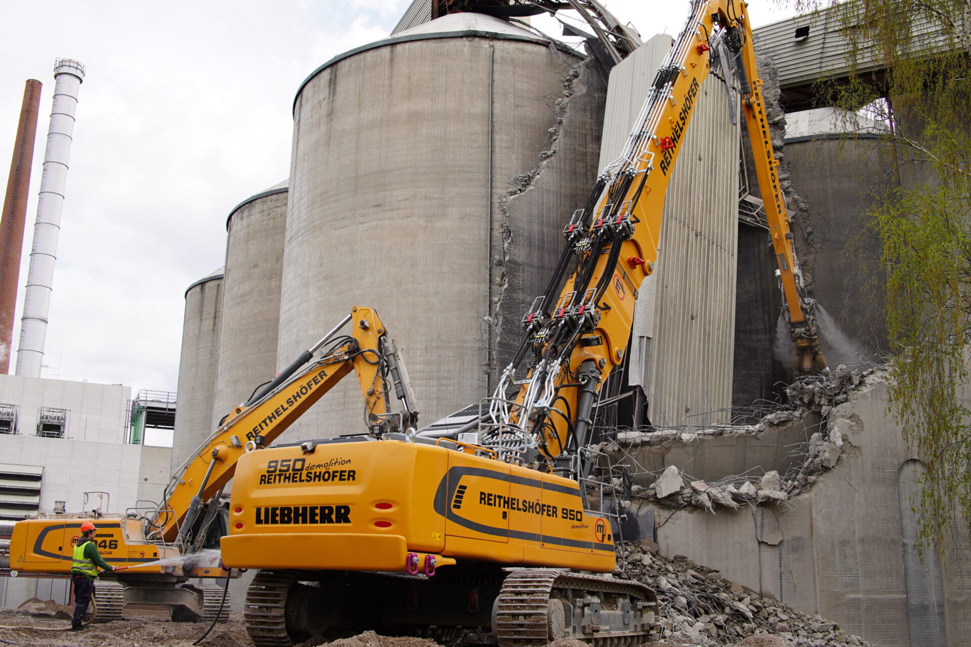 Auf dem Bild ist eine Baustelle zu sehen, auf der zwei große Bagger ein massives Betonbauwerk abreißen. Im Vordergrund steht ein Arbeiter in grüner Jacke nahe eines der Bagger. Im Hintergrund sind beschädigte zylindrische Betonstrukturen zu erkennen, vermutlich Silos.
