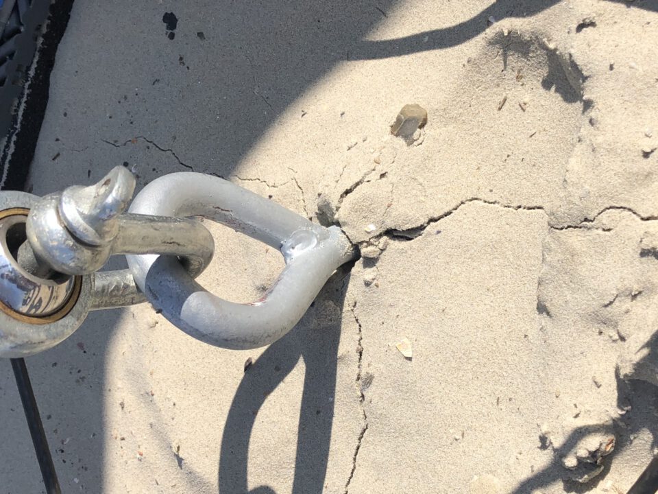 Das Bild zeigt einen Metallschäkel, der in eine rissige Betonfläche eingelassen ist. Der Schäkel ist mit einem weiteren Metallteil verbunden, möglicherweise Teil eines Ketten- oder Seilsystems. Um den Verankerungspunkt herum sind deutliche Risse im Sand zu sehen.