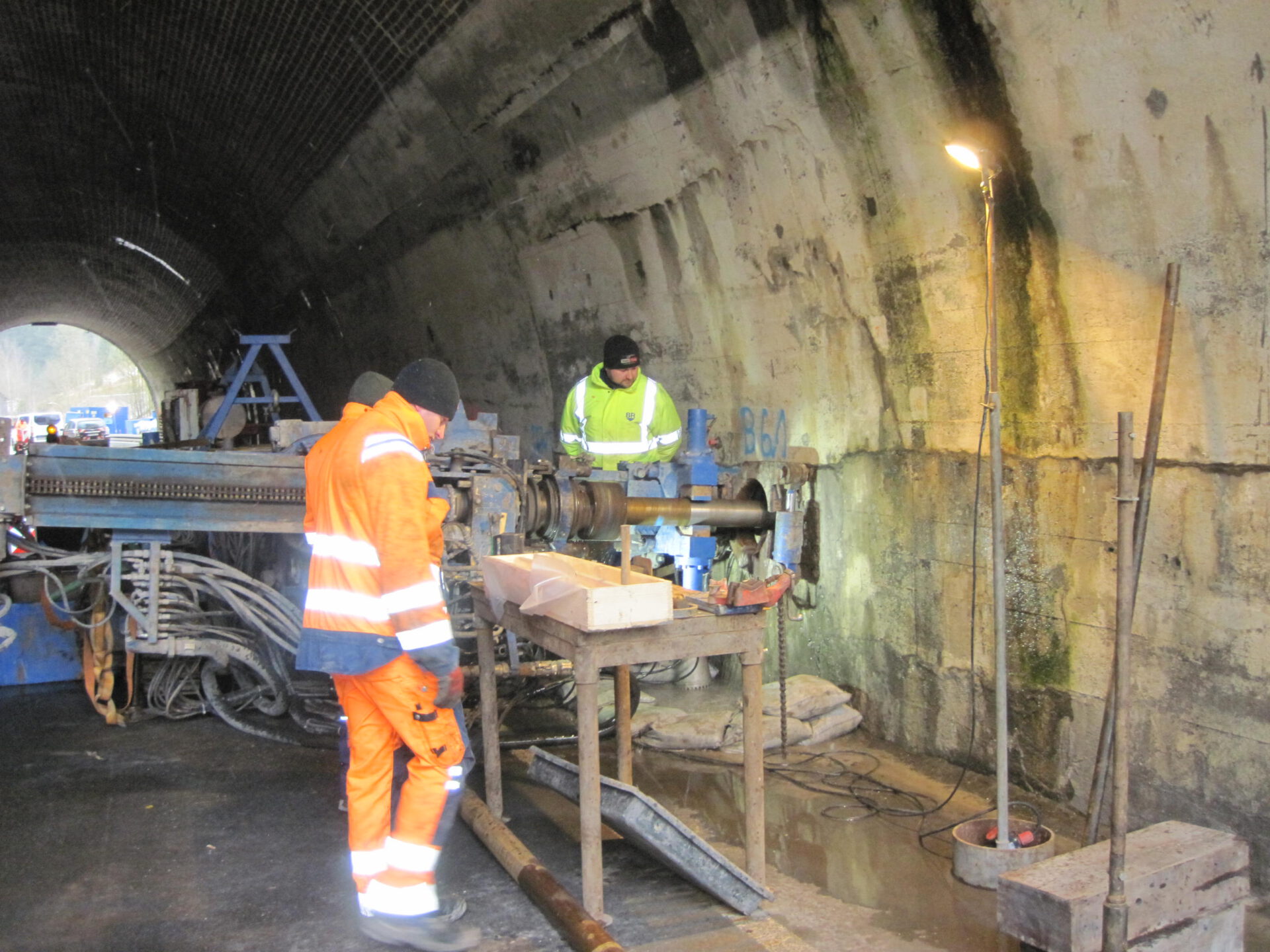 Das Bild zeigt zwei Arbeiter in einem Tunnel, einer in orangener und einer in gelber Warnkleidung. Sie bedienen Maschinen, vermutlich für Bohr- oder Wartungsarbeiten an der Tunnelwand. Um sie herum befinden sich Werkzeuge, Kabel, ein Arbeitstisch mit Holzklötzen und eine Stehlampe zur Ausleuchtung des Arbeitsbereichs.