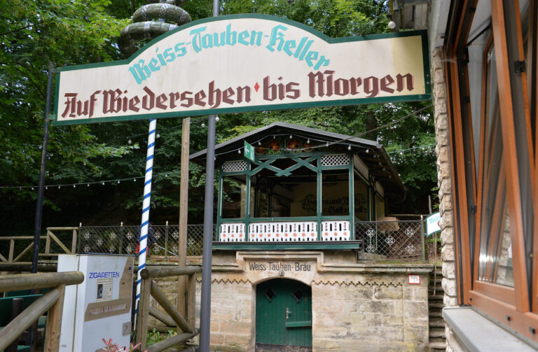 Auf dem Bild ist ein Braukeller zu sehen. Die Umgebung ist von Bäumen umgeben, mit Holzgeländern und einer kleinen Struktur mit grünem Eingang, die auf den Braukeller hinweist.