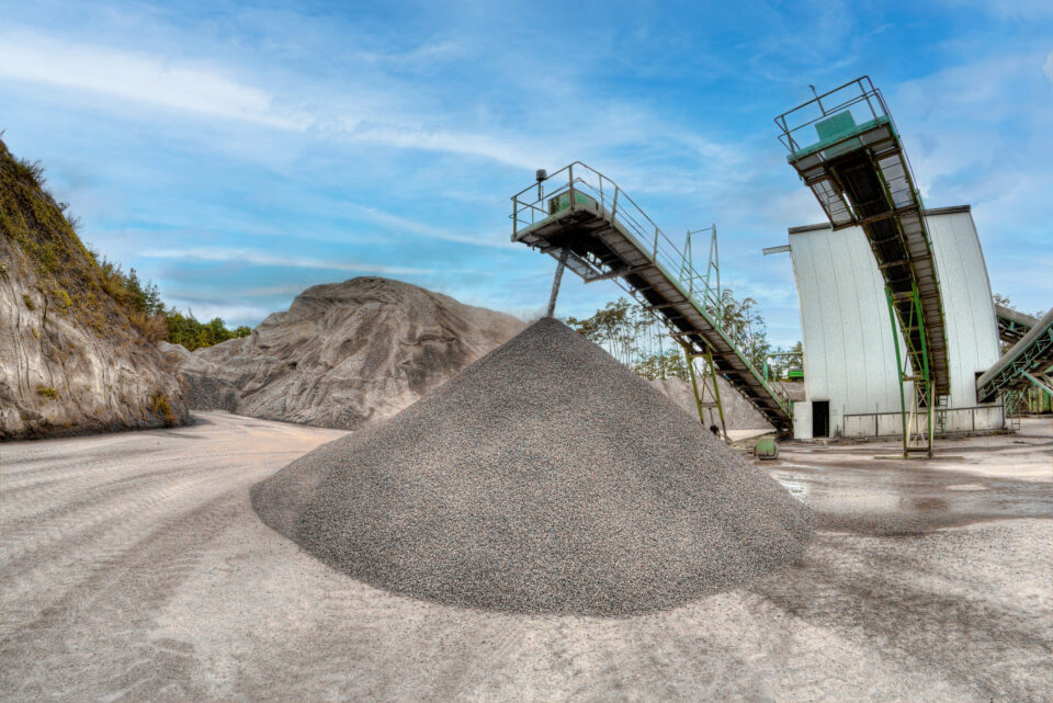 Das Bild zeigt ein Industriegelände mit einem großen Kies- oder Steinhaufen in der Mitte. Zwei erhöhte Förderbänder, gestützt von Metallkonstruktionen, verlaufen links und rechts und dienen dem Materialtransport. Im Hintergrund ist ein großer Erd- oder Felshügel zu sehen unter einem teils bewölkten Himmel.