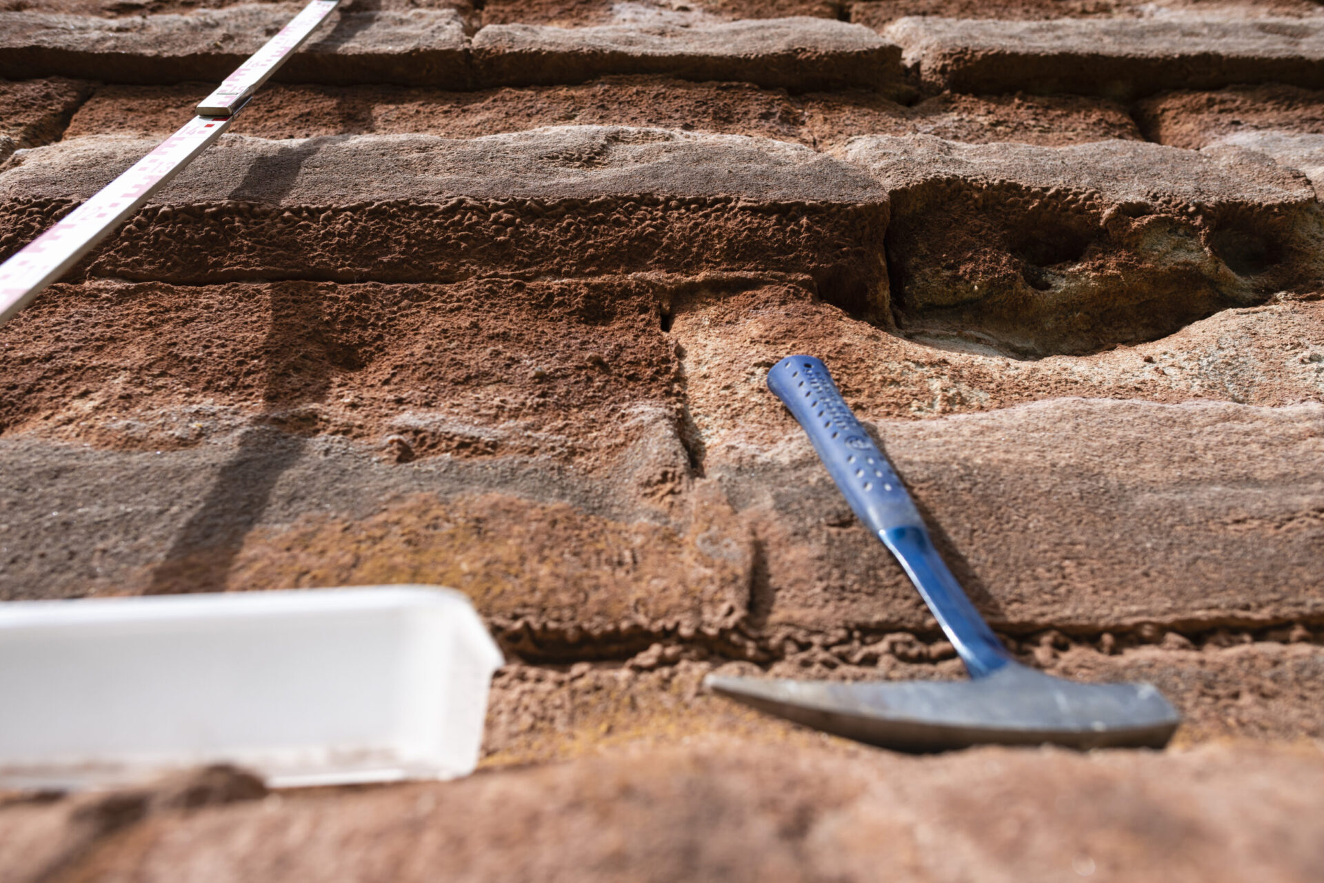 Das Bild zeigt eine Nahaufnahme einer Gesteinsoberfläche mit deutlich erkennbaren Schichten. Links ist ein Maßband senkrecht angebracht, um die Größenverhältnisse der Gesteinsschichten zu verdeutlichen. Rechts liegt ein Geologiehammer mit blauem Griff auf einer der Schichten, unten links befindet sich ein weißer Kunststoffbehälter.