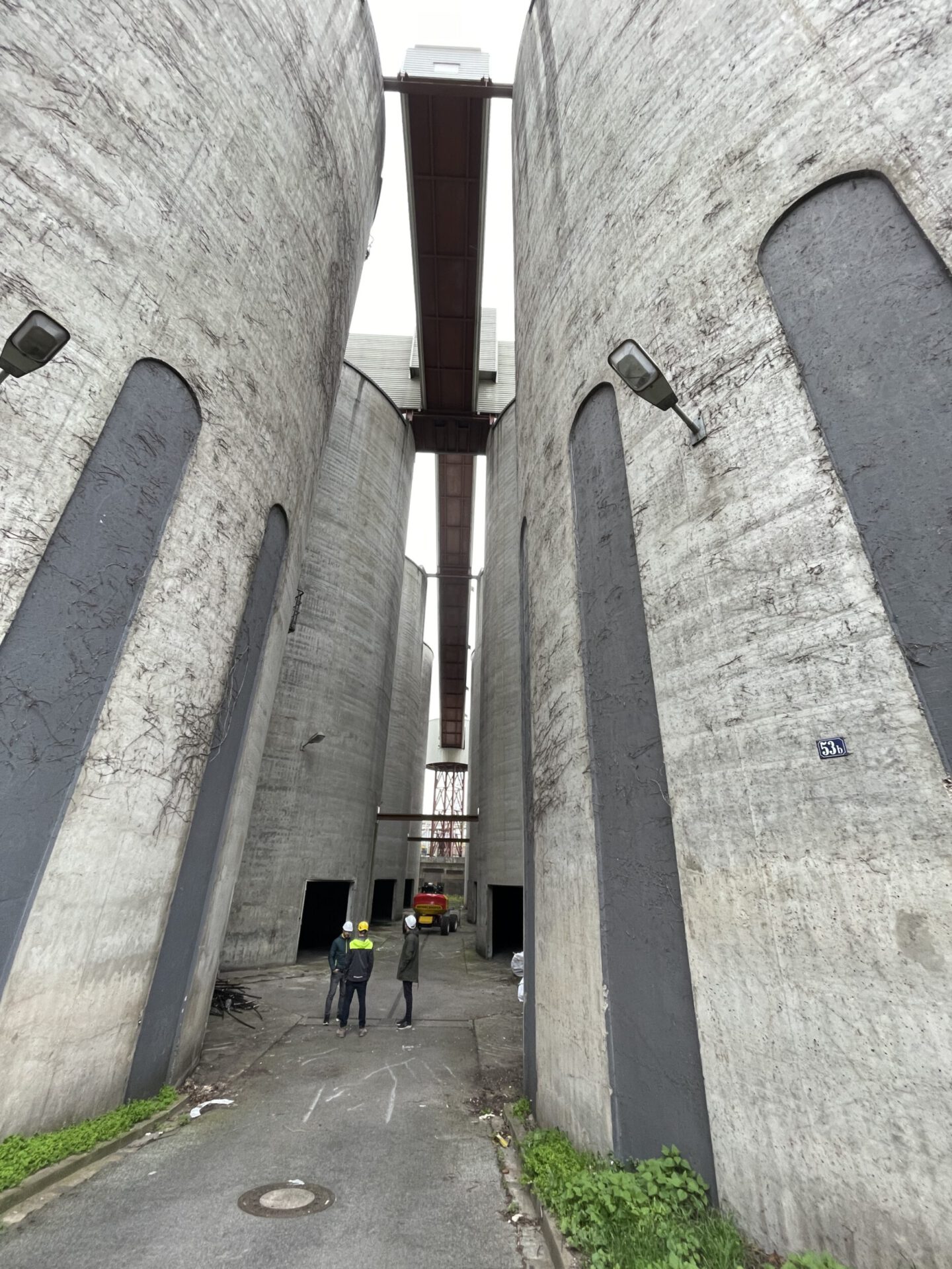 Auf dem Bild ist eine schmale Gasse zwischen großen, zylindrischen Betonbauten zu sehen. Zwei Personen stehen im Vordergrund, weiter hinten ist ein Fahrzeug erkennbar. Über den Bauwerken verlaufen Metallstege, und am Boden sind Pflastersteine sowie etwas Grün zu sehen.