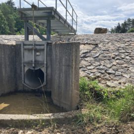 Das Bild zeigt eine Betonstruktur mit einem Metalltor oder -ventil, vermutlich Teil eines Wasserbauwerks wie eines Damms oder Überlaufs. Die Anlage ist in einen mit Steinen bedeckten Damm eingebettet, darüber verläuft ein Metallsteg. Die Umgebung ist leicht bewachsen und wirkt stellenweise schlammig.