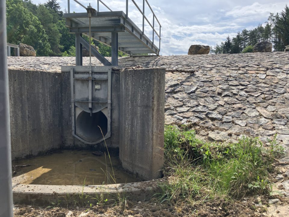 Das Bild zeigt eine Betonstruktur mit einem Metalltor oder -ventil, vermutlich Teil eines Wasserbauwerks wie eines Damms oder Überlaufs. Die Anlage ist in einen mit Steinen bedeckten Damm eingebettet, darüber verläuft ein Metallsteg. Die Umgebung ist leicht bewachsen und wirkt stellenweise schlammig.