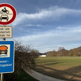 Das Bild zeigt eine ländliche Landschaft mit klarem Himmel und Hügeln im Hintergrund. Im Vordergrund stehen zwei Verkehrsschilder: eines verbietet die Durchfahrt für Motorräder und Autos, das andere weist auf ein Wasserschutzgebiet hin. Darunter befindet sich ein Zusatzschild, das land- und forstwirtschaftlichen Verkehr erlaubt.
